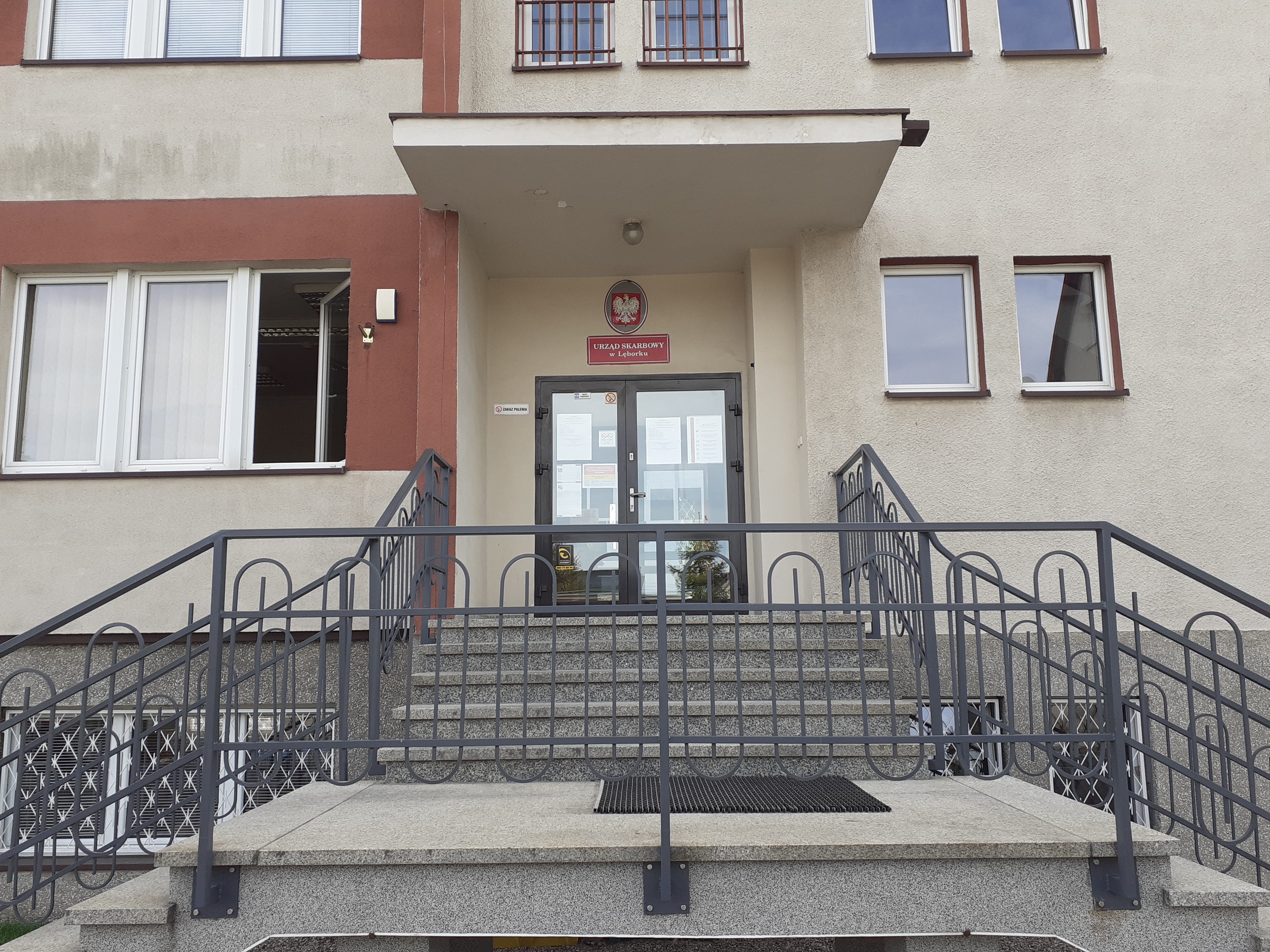 Wejście po schodach do budynku, w którym mieści się siedziba Urzędu Skarbowego w Lęborku. Szaro - brązowa elewacja