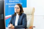 Minister Magdalena Rzeczkowska siedzi na fotelu przy biurku.
Z tyłu roll-up z logo krajowej Administracji Skarbowej.