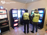 Funkcjonariusze Służby Celno-Skarbowej i Policji stoją przed automatami do gier hazardowych. W lewym górnym rogu znajduje się logo Krajowej Administracji Skarbowej.