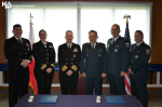 Pięciu mężczyzn  i kobieta w mundurach stoją przed stołem. Podpisują dokumenty. Z tyłu flaga Polski i Stanów Zjednoczonych