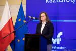 Kobieta w okularach stoi przed mikrofonem na tle niebieskiej ściany z logo Ministerstwa Finansów. Z boku po lewej stronie umieszczona jest flaga Polski i Unii Europejskiej.