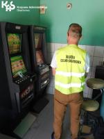 Funkcjonariusz Służby Celno-Skarbowej w żółtej kamizelce stoi przed automatami do gier hazardowych. W lewym górnym rogu logo KAS.