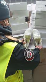 Funkcjonariuszka Służby Celno-Skarbowej trzyma w ręku dwa białe buty. W tle kartony z butami. W lewym górnym rogu logo Krajowej Administracji Skarbowej.