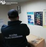 Funkcjonariusz Służby Celno-Skarbowej stoi przed biurkiem, na którym leżą białe koralowce, Na ścianie wisi kolorowy plakat, W lewym górnym rogu logo Krajowej Administracji Skarbowej.