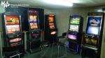 Ciemne pomieszczenie, w którym znajduje się kilka automatów do gier hazardowych. W lewym górnym rogu logo Krajowej Administracji Skarbowej.