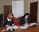 Dwie kobiety siedzą przy stole i podpisują dokumenty. W tle baner Krajowej Administracji Skarbowej.