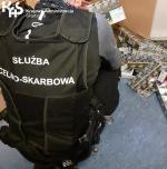 Funkcjonariusz Służby Celno-Skarbowej w czarnej kamizelce kuca na podłodze. Z boku po prawej stronie porozrzucane kartony z papierosami. W lewym górnym rogu logo Krajowej Administracji Skarbowej.