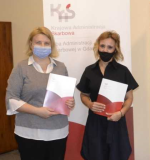 Dwie kobiety stoją w pomieszczeniu, w rękach trzymają dokumenty, za nimi baner KAS, 
