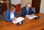Kobieta i mężczyzna siedzą przy biurku. Podpisują dokumenty. W lewym górnym rogu logo Krajowej Administracji Skarbowej.