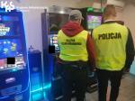 Funkcjonariusz Służby Celno-Skarbowej i Policji w zółtych kamizelkach w salonie z nielegalnymi automatami do gier hazardowych.Po lewej stronie ustawione automaty do gier. W lewym górnym rogu logo Krajowej Administracji Skarbowej.
