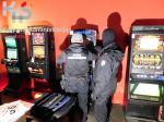 Duże pomieszczenie w kolorze czerwonym. Dwóch funkcjonariuszy Służby Celno-Skarbowej stoi przy automatach do gier hazardowych. W lewym górnym rogu logo Krajowej Administracji Skarbowej.