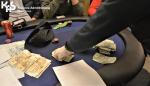 Stół do pokera, na którym widać między innymi dłonie, pieniądze i żetony. W lewym górnym rogu logo KAS. 
