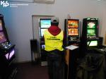 Jasne pomieszczenie, w którym znajdują się automaty do gier hazardowych.Funkcjonariusz Służby Celno-Skarbowej stoi przodem przed jednym z automatów. W lewym górnym rogu logo Krajowej Administracji Skarbowej.