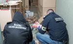 Funkcjonariusze Służby Celno-Skarbowej oraz Straży Granicznej klęczą przy kartonach z papierosami. W tle poukładane kartony z papierosami.
W lewym górnym rogu logo Krajowej Administracji Skarbowej.