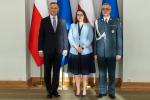 Nominacja generalska. W sali Pałacu Prezydenckiego na tle flag Polski i Unii Europejskiej stoją: Prezydent  Rzeczypospolitej Polskiej, Minister Finansów oraz nowo mianowany generał.