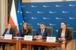 Przy stole siedzą cztery osoby: trzy kobiety i jeden mężczyzna. w tle niebieski baner Krajowej Administracji Skarbowej. Po lewej stronie stoi flaga Polski i Unii Europejskiej.