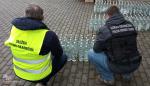 Funkcjonariusze Służby Celno-Skarbowej liczą butelki z alkoholem.
Butelki ustawione na ziemi.